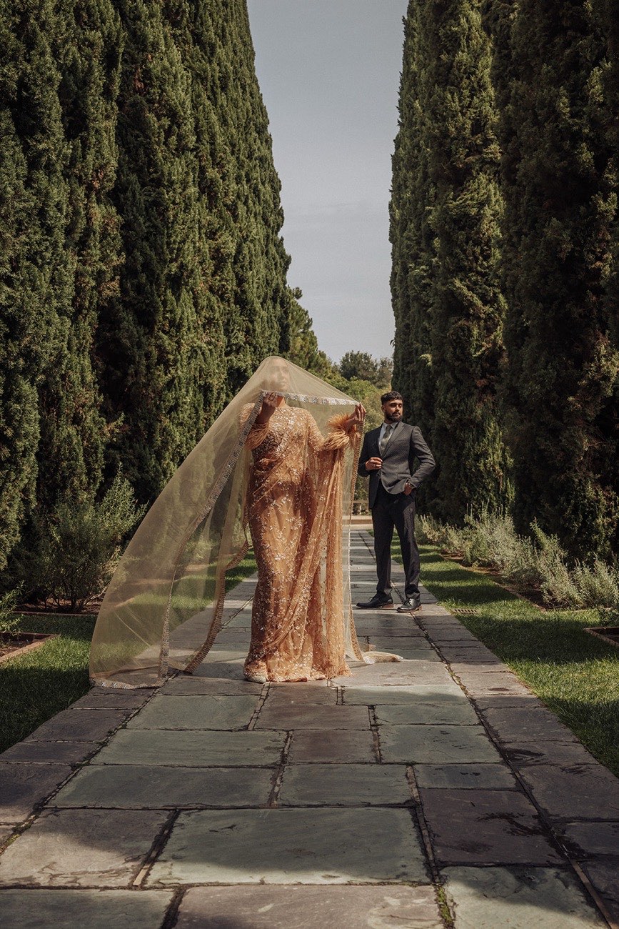 A South Asian bride under a flowing gold veil beside the groom on a stone path between towering cypress trees