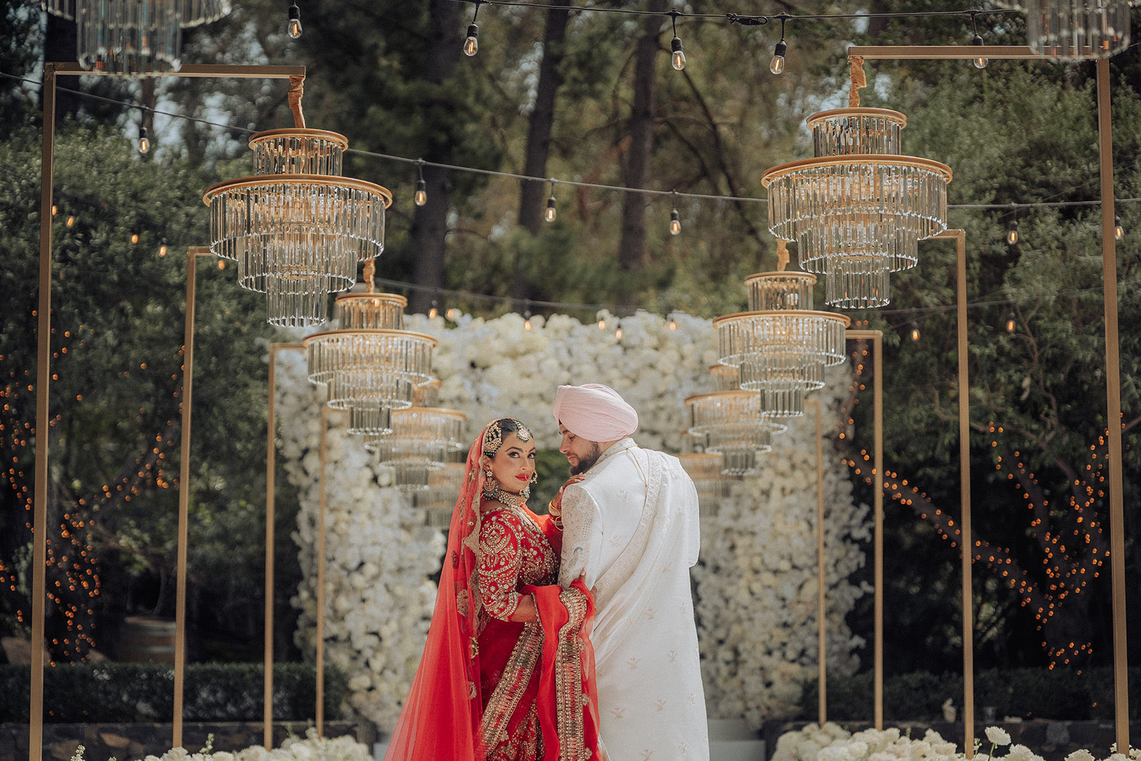 Gunjan and Vikram destination Sikh wedding at Calamigos Ranch, Malibu — the couple beneath tiered crystal chandeliers and a white floral wall, the bride in a red lehenga and the groom in an ivory sherwani and turban