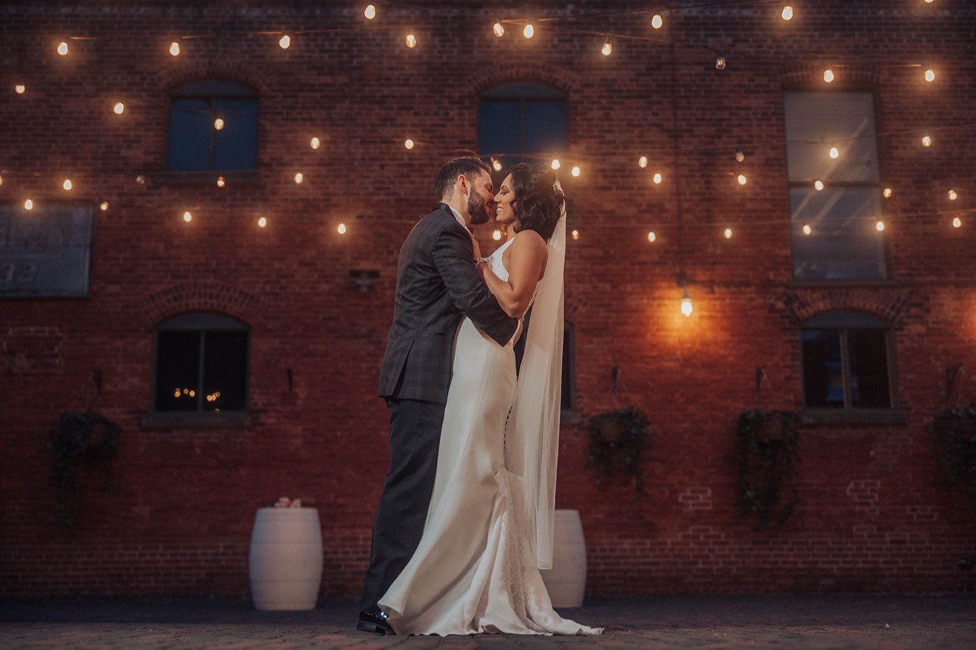 Haramrit and Ethan wedding — the couple embracing under string lights against a brick wall in Toronto's Distillery District