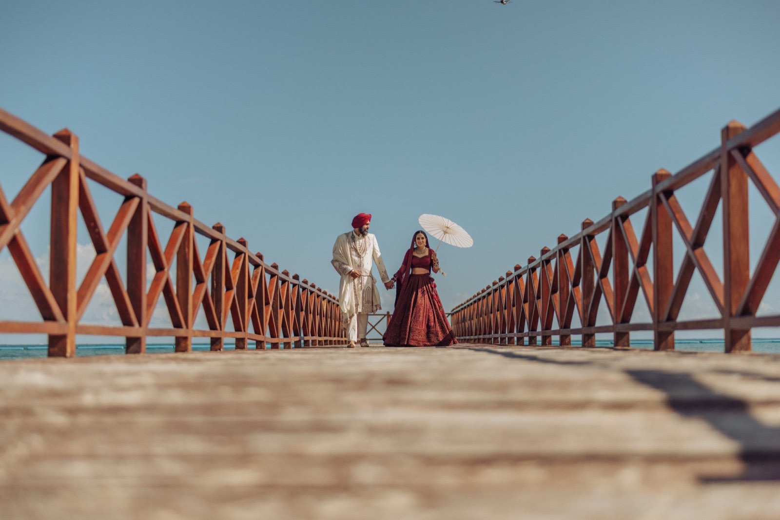Jeet and Ronvir destination Sikh wedding at Moon Palace, Cancun — the couple walking a wooden pier over turquoise ocean, the groom in an ivory sherwani and red turban and the bride in a maroon lehenga with a paper parasol