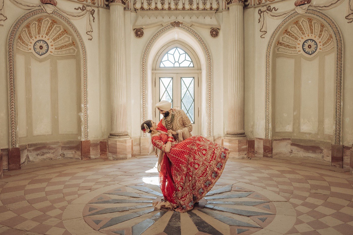 Laura and Simar destination Sikh wedding in Germany — the bride in a red bridal lehenga and the groom in a turban and sherwani in an ornate domed hall