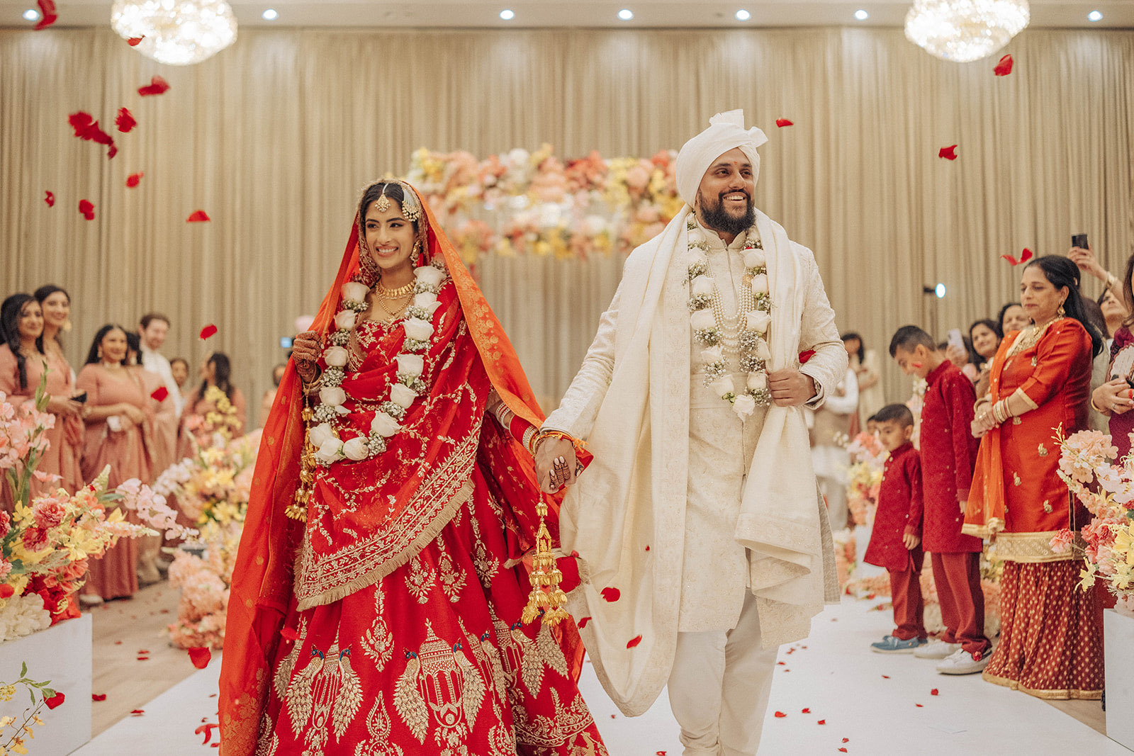 Manpreet and Vinay Sikh and Hindu wedding — the couple walking through a shower of rose petals, the bride in a red lehenga and the groom in an ivory sherwani and turban