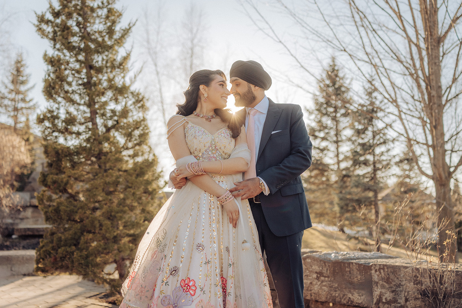 Moni and Bhupinder engagement — the couple embracing at golden hour among evergreens at the Royal Ambassador in Caledon, the bride in a cream floral lehenga and the groom in a turban and navy suit