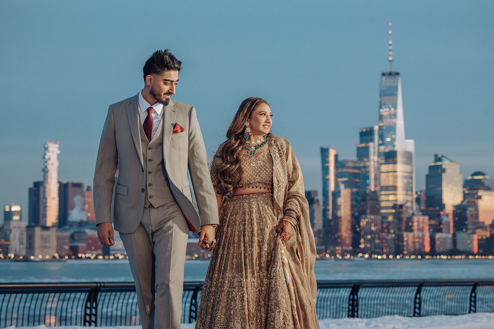 Natalya and Amritpal engagement — the couple walking the Hoboken waterfront at golden hour with the Manhattan skyline and One World Trade Center behind them, the bride in a gold lehenga and the groom in a grey suit