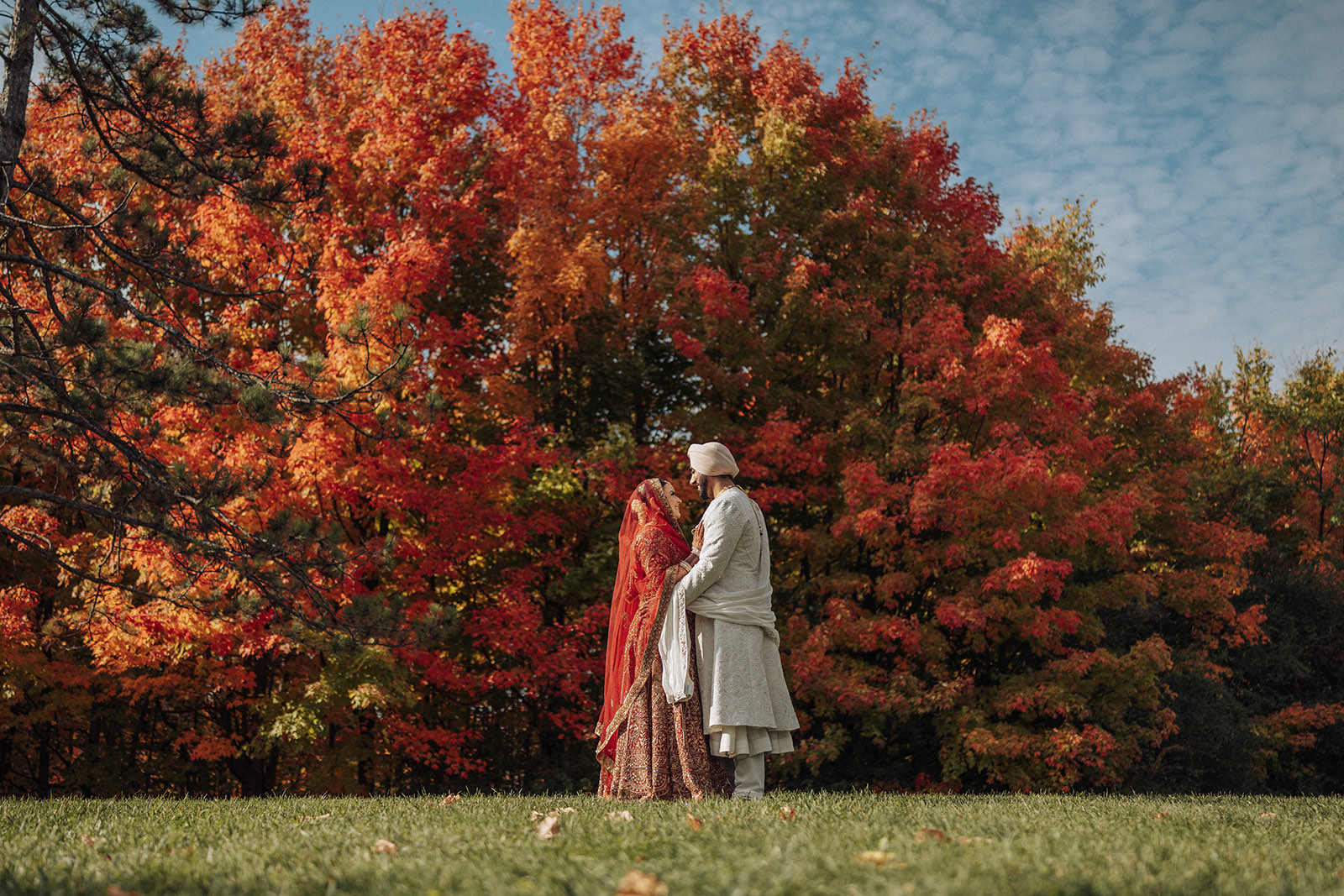 Navjot and Rajvinder Sikh wedding portraits at White Spruce Park, Brampton — the bride in a red lengha and veil and the groom in a cream sherwani and turban against vivid red and orange fall foliage