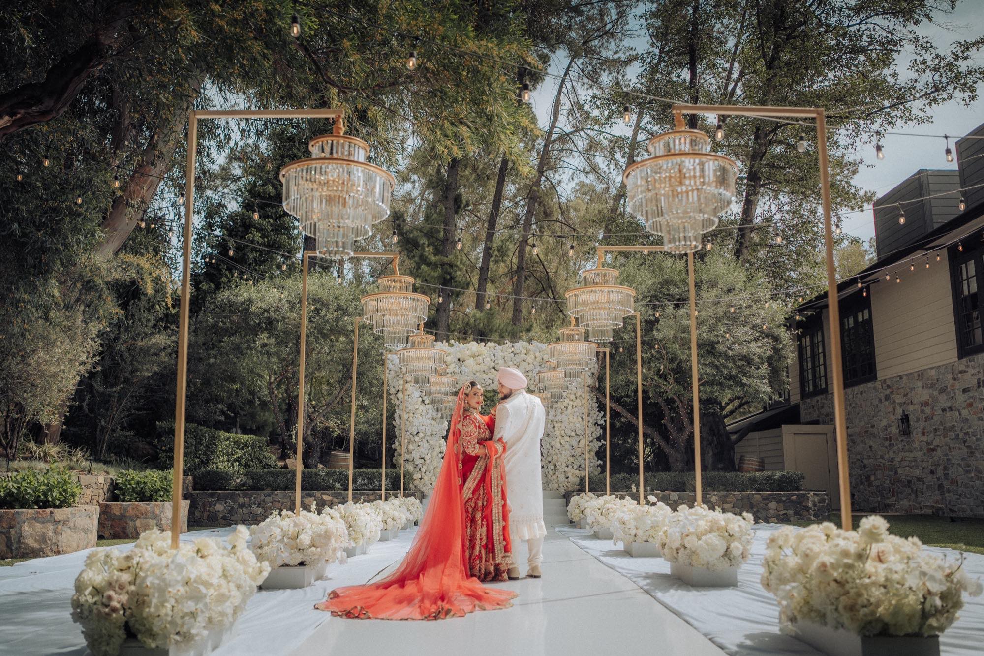 A Sikh bride in a red bridal lehenga with a long trailing dupatta and the groom in a white sherwani and turban at an outdoor wedding, framed by tiered crystal chandeliers, a white floral wall and rows of white roses