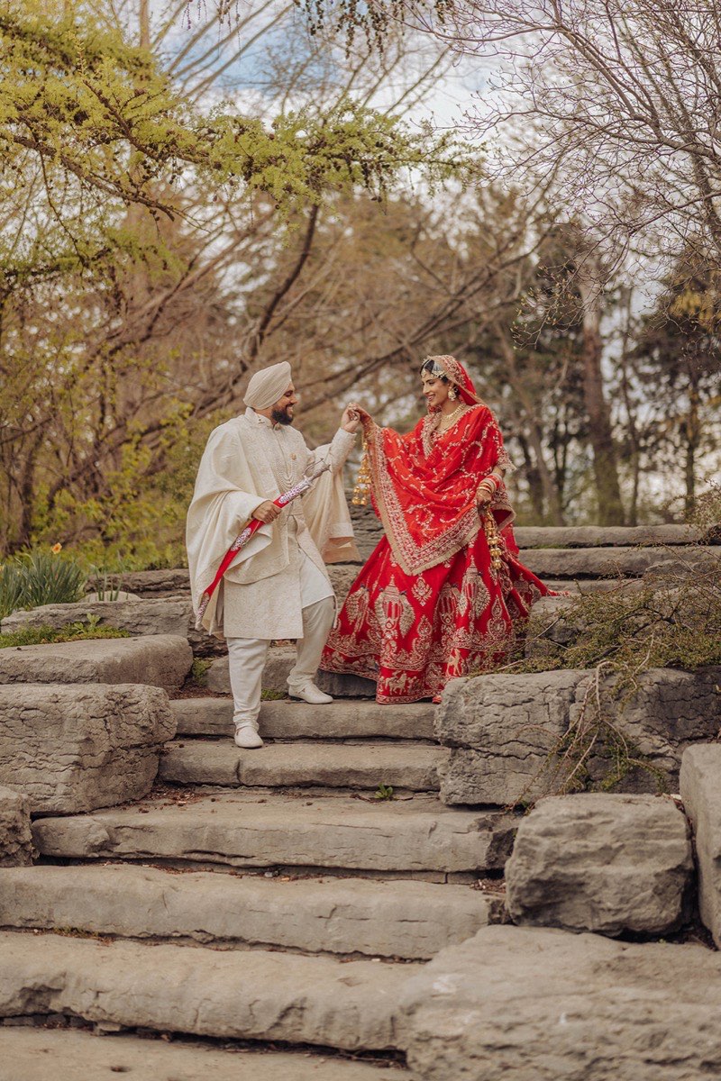 A Sikh bride in a red bridal lehenga and groom in a white sherwani on the stone steps at Milliken Park in Scarborough