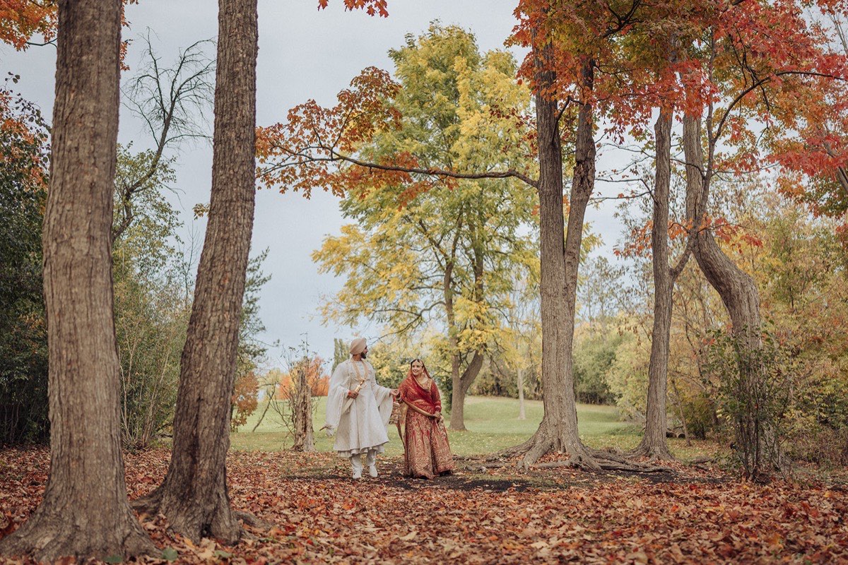 A Sikh couple in wedding attire walking among autumn trees and fallen leaves at White Spruce Park in Brampton