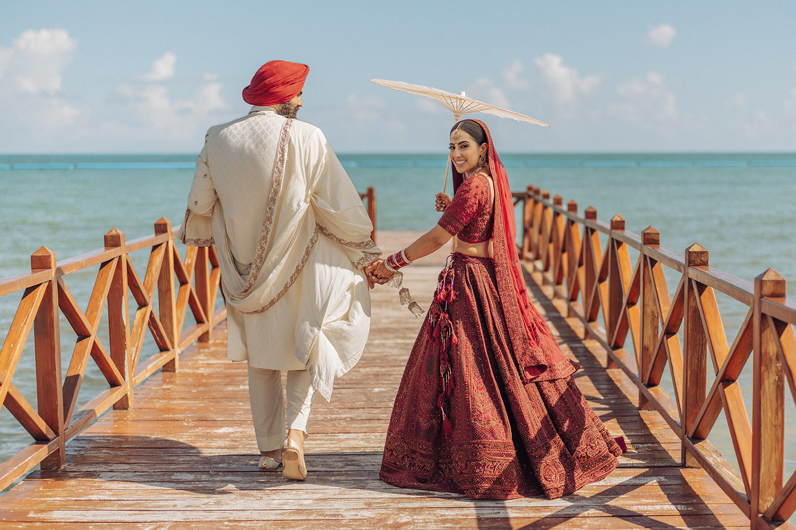 A Sikh bride in a red lehenga holding a parasol and the groom in an ivory sherwani and red turban walking along an ocean pier at a destination wedding